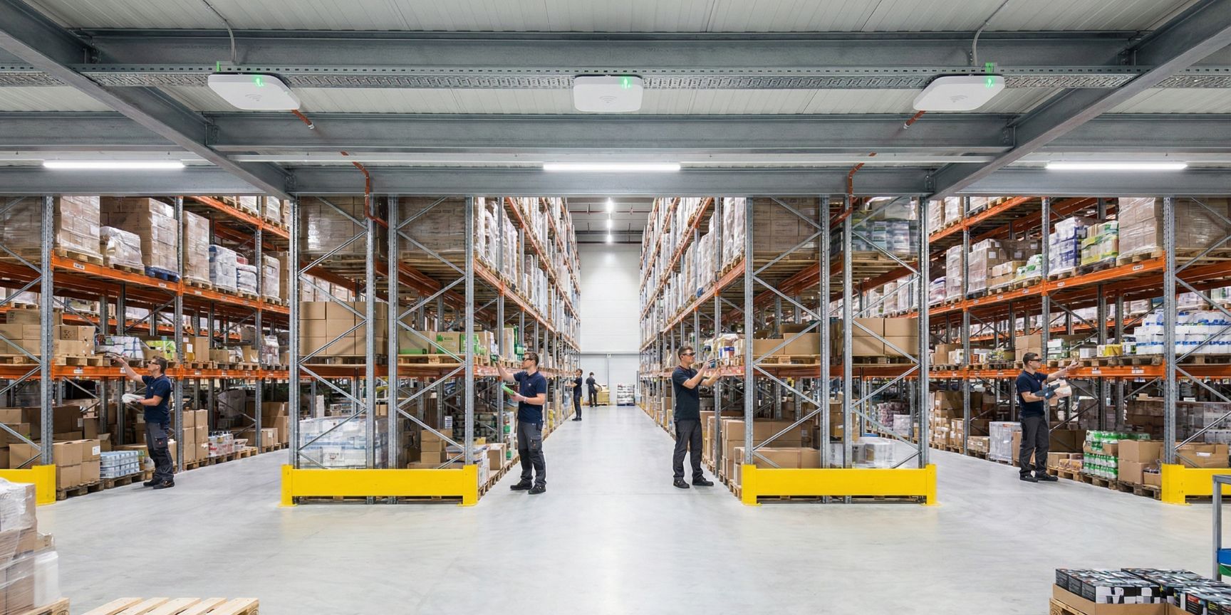 Wide-angle view of a modern high-bay warehouse with several employees wearing data glasses for order picking. Several WLAN access points are clearly installed on the hall ceiling for comprehensive illumination